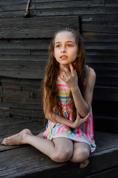 Pensive little child with long hair resting on wooden deck, looking aside while dreaming about something. - Photo, Image