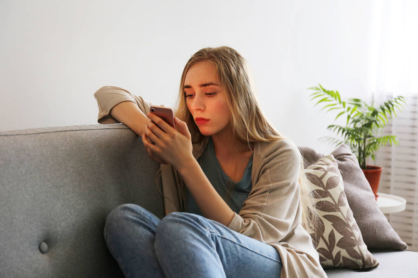 Portrait of beautiful young woman with depressed facial expression sitting on grey textile couch holding her phone. Cyber bullying victim concept. Sad female in her room. Background, copy space. - Fotografie, Obrázek