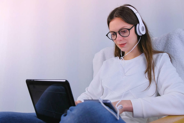 Woman in glasses and headphones is working on laptop sitting in armchair. - Fotó, kép