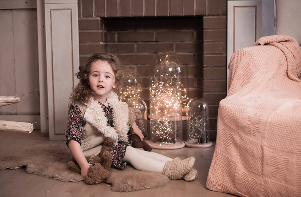 The baby girl in a fur vest sits near a fireplace. A winter portrait - Foto, Bild