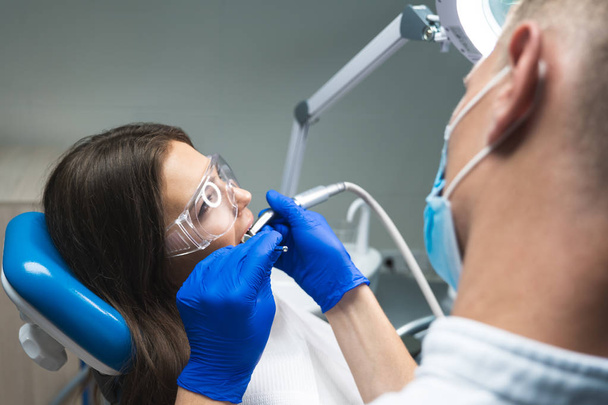 dentist in medical mask filling the patients root canal while she is lying on dental chair wearing safety glasses - Foto, Imagen