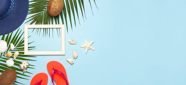 Summer fashion flat lay. Blue hat coral flip flops tropical palm leaves sunscreen white photo frame coconut seashells starfish on blue background. Top view copy space. Creative vacation background - Photo, Image