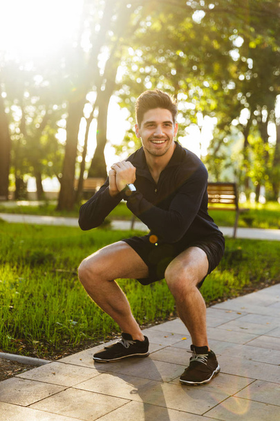Cheerful young sports fitness man standing in green park nature make sport exercises for legs squats. - Photo, image