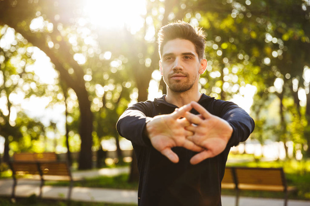 Serious young sports fitness man standing in green park nature make stretching exercises for arms. - 写真・画像