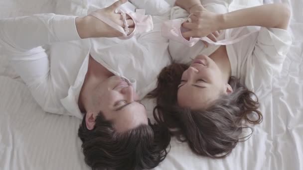 Happy couple lying in bed playing with pink childrens pointe and hugging close-up. Young family waiting for a baby girl. Tender relationship concept, people in love. Top view. Slow motion. - Filmagem, Vídeo