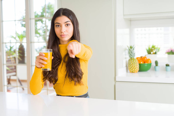 Young woman drinking a glass of fresh orange juice pointing with finger to the camera and to you, hand sign, positive and confident gesture from the front - Foto, Imagen
