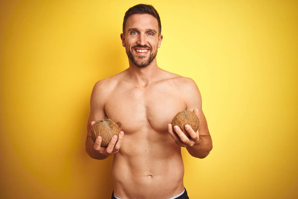 Homem sem camisa bonito jovem segurando coco tropical exótico sobre fundo amarelo isolado com um rosto feliz de pé e sorrindo com um sorriso confiante mostrando dentes
 - Foto, Imagem