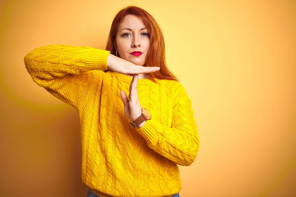 schöne rothaarige Frau im Winterpullover, die vor isoliertem gelben Hintergrund steht und eine Auszeit macht, Geste mit Händen, frustriertem und ernstem Gesicht - Foto, Bild