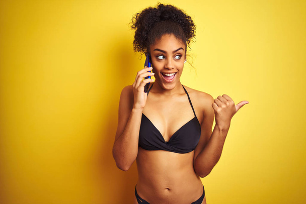 Afro american woman wearing bikini talking using smartphone over isolated yellow background pointing and showing with thumb up to the side with happy face smiling - Foto, afbeelding