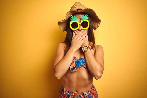 Mujer de vacaciones con bikini y gafas de sol de piña sobre fondo amarillo aislado impactó cubriendo la boca con las manos por error. Concepto secreto
. - Foto, Imagen