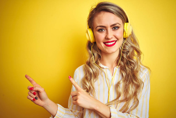 Молодая красивая женщина слушает музыку с помощью наушников на желтом изолированном фоне улыбаясь и глядя на камеру указывая двумя руками и пальцами в сторону
. - Фото, изображение
