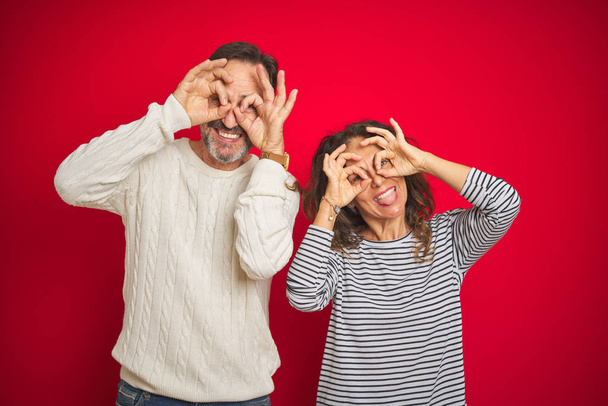 Hermosa pareja de mediana edad que usa suéter de invierno sobre un fondo rojo aislado haciendo un gesto aceptable como binoculares sacando la lengua, ojos mirando a través de los dedos. Expresión loca
. - Foto, imagen