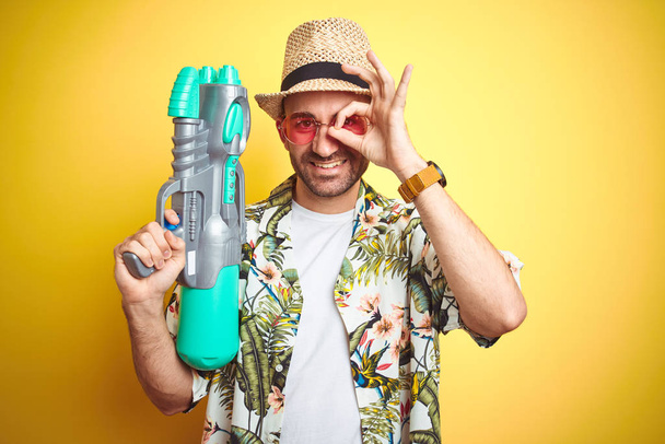 Молодой человек в гавайских цветах рубашку с водяным пистолетом на желтом изолированном фоне со счастливым лицом улыбается делает хорошо знак с рукой на глазу, глядя сквозь пальцы
 - Фото, изображение
