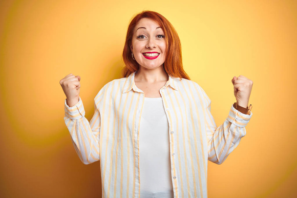 Beautiful redhead woman wearing striped shirt standing over isolated yellow background celebrating surprised and amazed for success with arms raised and open eyes. Winner concept. - Photo, Image