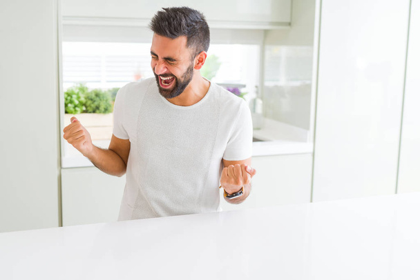 T-shirt blanc décontracté homme hispanique beau à la maison très heureux et excité faisant geste gagnant avec les bras levés, souriant et criant pour le succès. Concept de célébration
. - Photo, image
