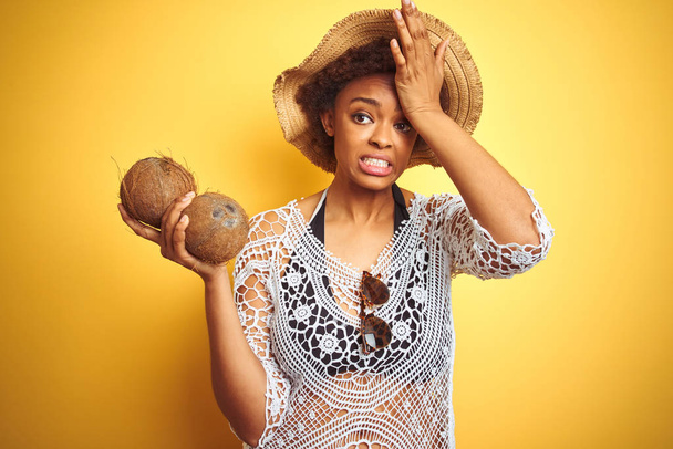 Молодая африканская американка с афроволосами, держащая кокос на жёлтом изолированном фоне, напряжённая рукой на голове, шокированная стыдом и удивлением, сердитая и разочарованная. Страх и разочарование по ошибке
. - Фото, изображение
