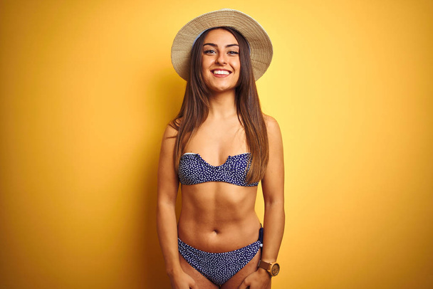 Young beautiful woman on vacation wearing bikini and hat over isolated yellow background with a happy and cool smile on face. Lucky person. - Photo, Image
