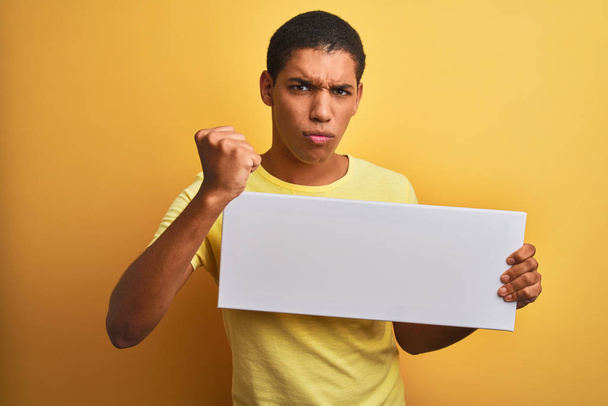 Young handsome arab man holding banner standing over isolated yellow background annoyed and frustrated shouting with anger, crazy and yelling with raised hand, anger concept - Foto, afbeelding