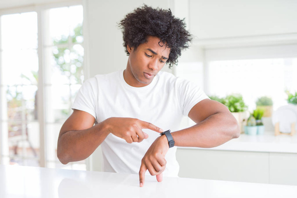 Hombre afroamericano joven con camiseta blanca casual sentado en casa Comprobación de la hora en el reloj de pulsera, relajado y confiado
 - Foto, Imagen