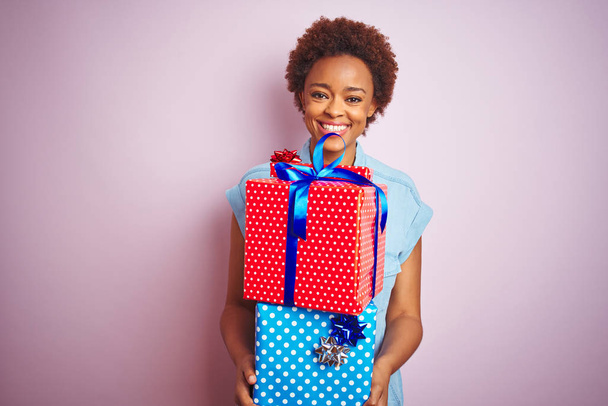 ピンクの孤立した背景の上に誕生日プレゼントを持つアフリカ系アメリカ人女性は、歯を見せる自信のある笑顔で立ち上がり、微笑んでいる - 写真・画像