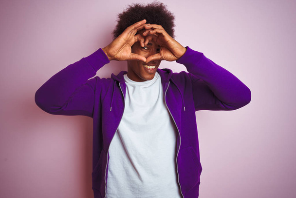 Jovem afro-americano vestindo camisola roxa em pé sobre fundo rosa isolado Fazendo forma de coração com a mão e os dedos sorrindo olhando através do sinal
 - Foto, Imagem