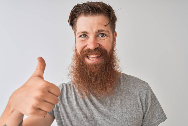 junger rothaariger irischer Mann trägt T-Shirt, das über isoliertem grauem Hintergrund steht, glücklich mit breitem Lächeln, das ok Zeichen macht, Daumen hoch mit Fingern, hervorragendes Zeichen - Foto, Bild