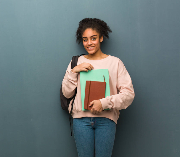 Молодая студентка черная женщина, скрещивающая руки, улыбающаяся и расслабленная. Она держит книги.
. - Фото, изображение