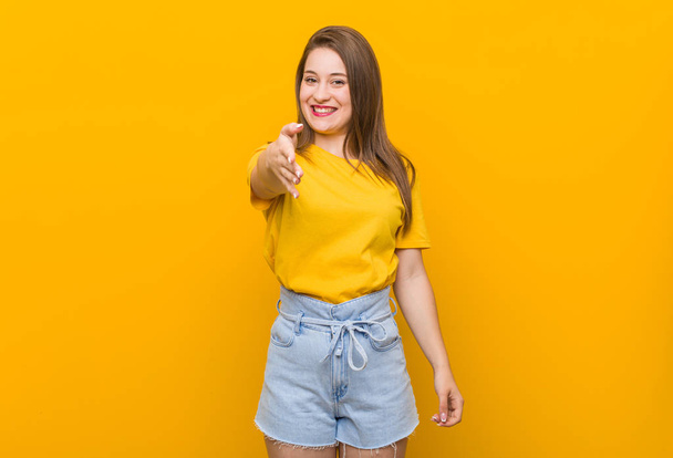 Młoda kobieta Nastolatka ubrana w żółtą koszulę rozciągającą rękę w aparacie w gest pozdrowienia. - Zdjęcie, obraz