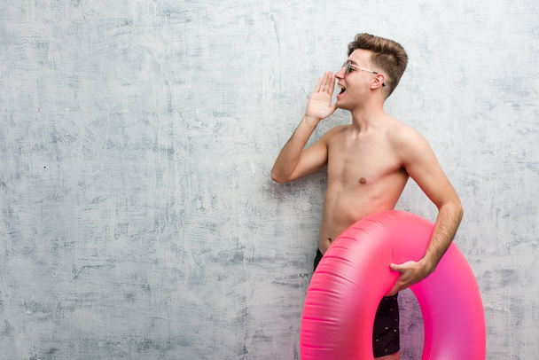 Young man holding a pink inflatable donut wearing a swimsuit shouting and holding palm near opened mouth. - Foto, afbeelding