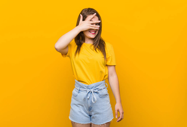Young woman teenager wearing a yellow shirt blink at the camera through fingers, embarrassed covering face. - Photo, Image