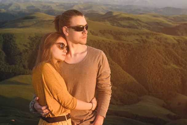 Portrait of a romantic young hipster couple hugging. They stand in an embrace in nature high in the mountains against the backdrop of a mountain green valley and clouds. - Foto, Imagem