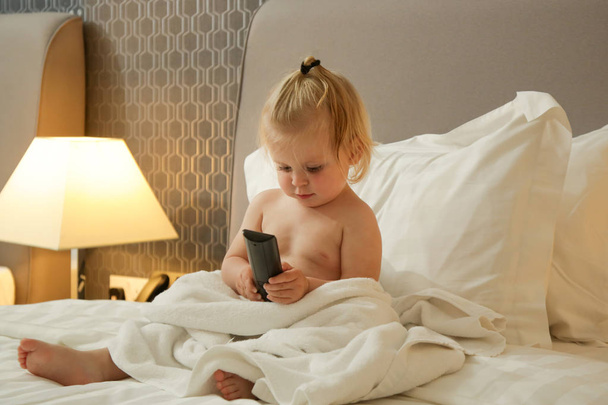 Χαριτωμένο κορίτσι νήπιο κάθεται στο κρεβάτι και κρατώντας τηλεχειριστήριο TV - Φωτογραφία, εικόνα
