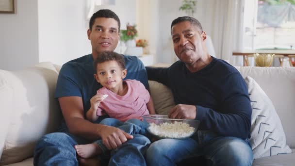 Three generation male family group sitting on a sofa eating popcorn and watching TV together - Imágenes, Vídeo