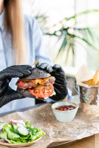 Young woman in blue shirt and black gloves eating delicious juicy hamburger with beef chop. Burgers in the cafe with salad and fries, girl eating fast food. - Photo, Image