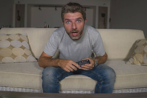 young attractive and relaxed man at living room sofa couch watching television in surprise and show holding remote enjoying thriller movie or mystery episode - Foto, Bild
