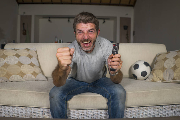 30s or 40s football fan man watching soccer game celebrating his team scoring goal crazy happy screaming cheering his team sitting at home living room couch gesturing - Fotó, kép