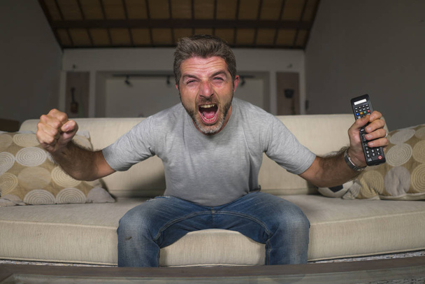 30s or 40s football fan man watching soccer game celebrating his team scoring goal crazy happy screaming cheering his team sitting at home living room couch gesturing - Foto, Bild