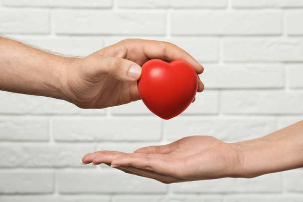Man giving red heart to woman at white brick wall, closeup. Donation concept - Foto, imagen