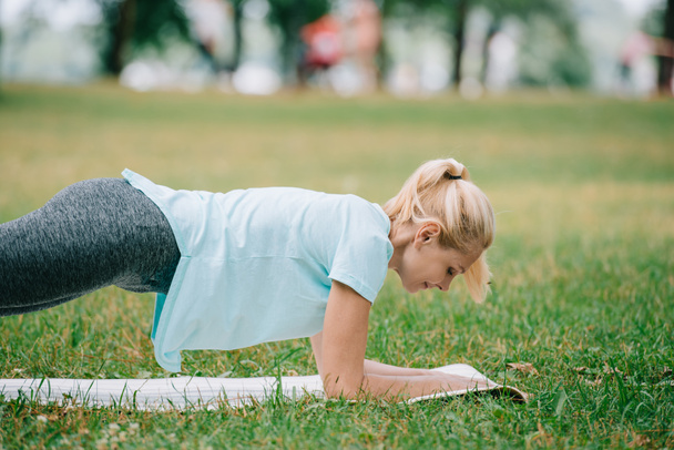 ελκυστική ώριμη γυναίκα κάνει άσκηση σανίδα, ενώ racticing γιόγκα σε πράσινο γκαζόν στο πάρκο - Φωτογραφία, εικόνα