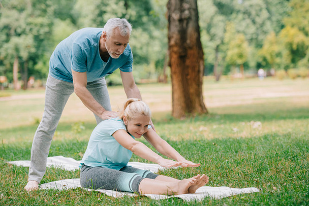 красивый зрелый мужчина помогает женщине практиковать йогу на лужайке в парке
 - Фото, изображение
