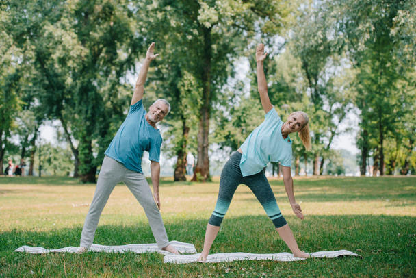 smiling mature man and woman practicing warrior yoga poses while standing on yoga mats in park - Foto, Imagen