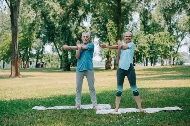 positive, mature man and woman practicing yoga while standing on yoga mats in park - Fotografie, Obrázek