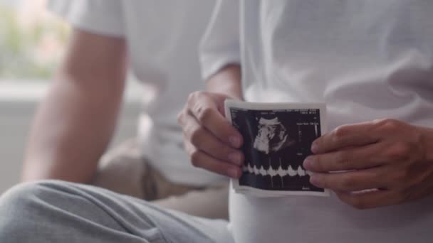 Young Asian Pregnant couple show and looking ultrasound photo baby in belly. Mom and Dad feeling happy smiling peaceful while take care child lying on sofa in living room at home concept. Slow motion. - Materiał filmowy, wideo