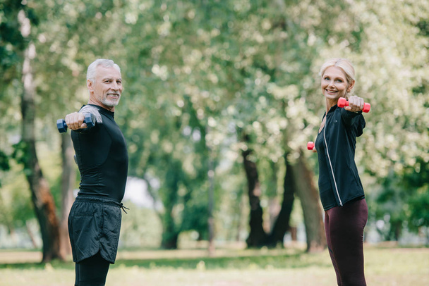 cheerful, mature sportsman and sportswoman training with dumbbells in park and smiling at camera - Photo, Image