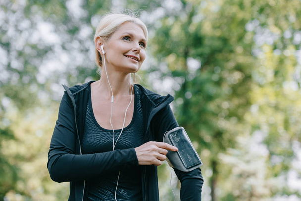 smiling mature sportswoman looking away while listening music in earphones - Photo, image