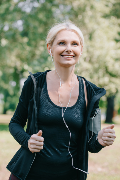 cheerful mature sportswoman jogging in park and listening music in earphones - Photo, Image