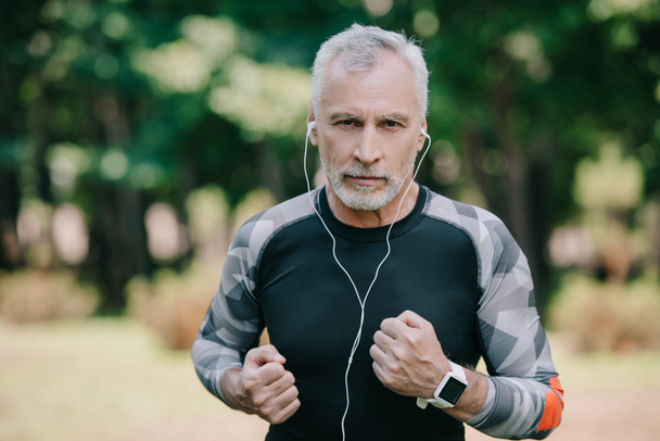 confident mature sportsman listening music in earphones while jogging in park - Photo, Image