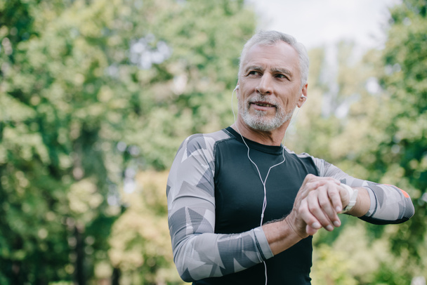 smiling mature sportsman touching fitness tracker while listening music in earphones - Photo, Image