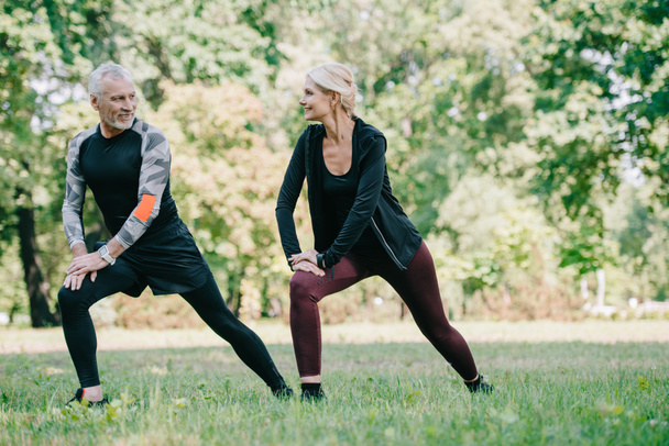 smiling, mature sportsman and sportswoman doing lunges exercises and looking at each other in park  - Photo, Image