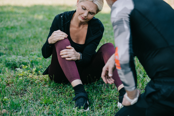 mature sportswoman sitting on lawn and touching injured leg near sportsman - Fotografie, Obrázek